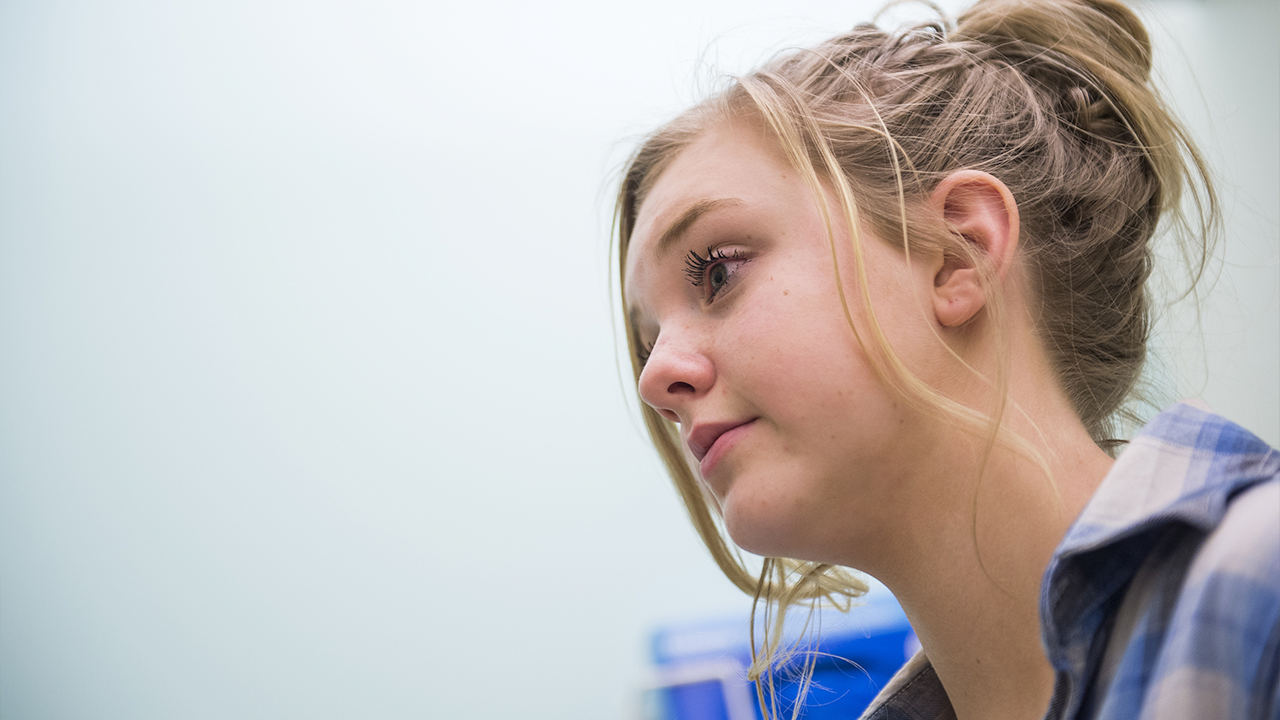 A close-up of a teenage girl with blonde hair in a bun and wearing a blue plaid shirt.