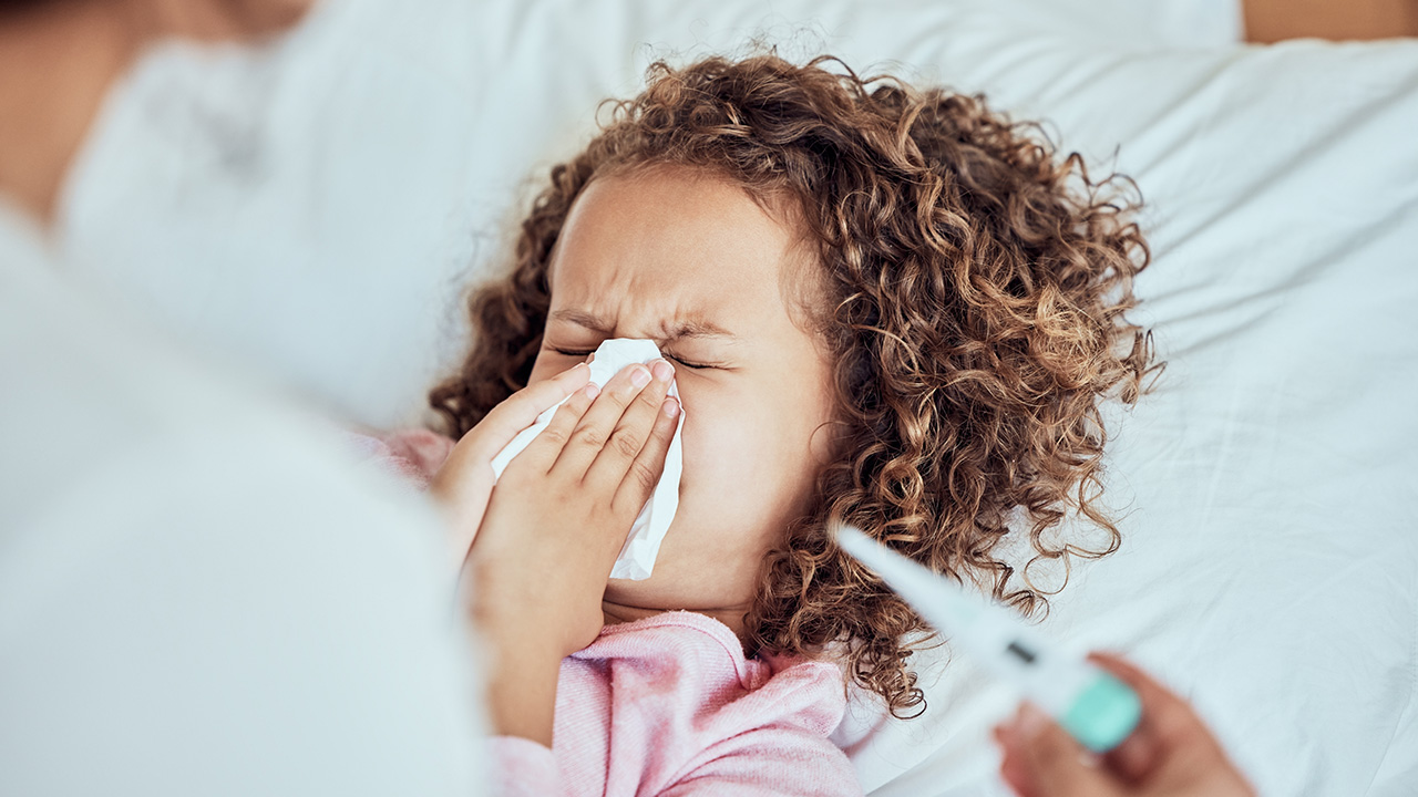 A young child sneezes into a tissue with their eyes shut while a caregiver holds a thermometer.