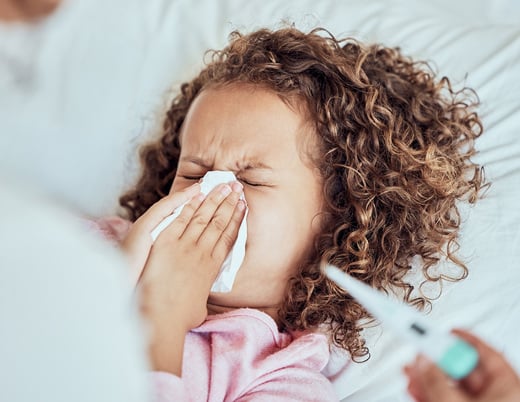 A young child sneezes into a tissue with their eyes shut while a caregiver holds a thermometer.