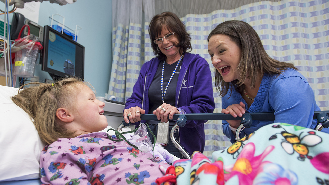 A girl lies in a hospital bed while her mom and another woman stand by her bed.