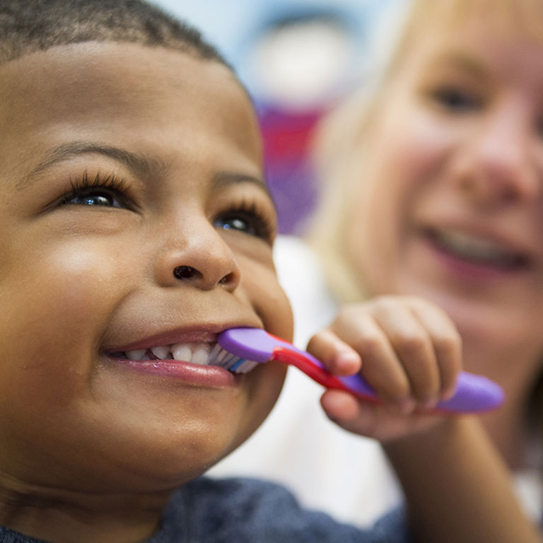 A young boy wearing a blue shirt smiles and brushes his teeth with a purple and pink toothbrush while a doctor with long blonde hair watches in the background.