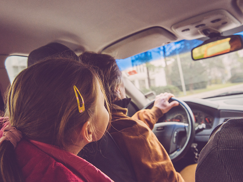 A girl wearing a pink winter coat, and her brown hair in a ponytail, sits in the back seat while a man in a brown coat drives the car.