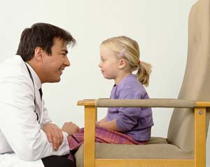 A doctor with brown hair and white lab coat squats down to eye level of a girl with blonde hair and wearing a purple shirt sitting in a beige chair.