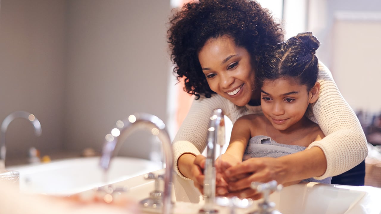 A woman wraps her arms around a young girl and helps her wash her hands over a sink.