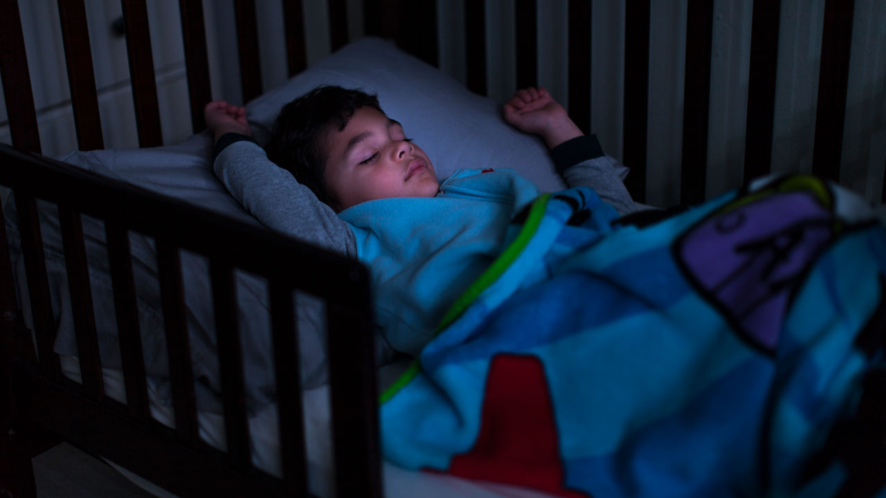 A toddler sleeps in a small bed with his arms up by his head.