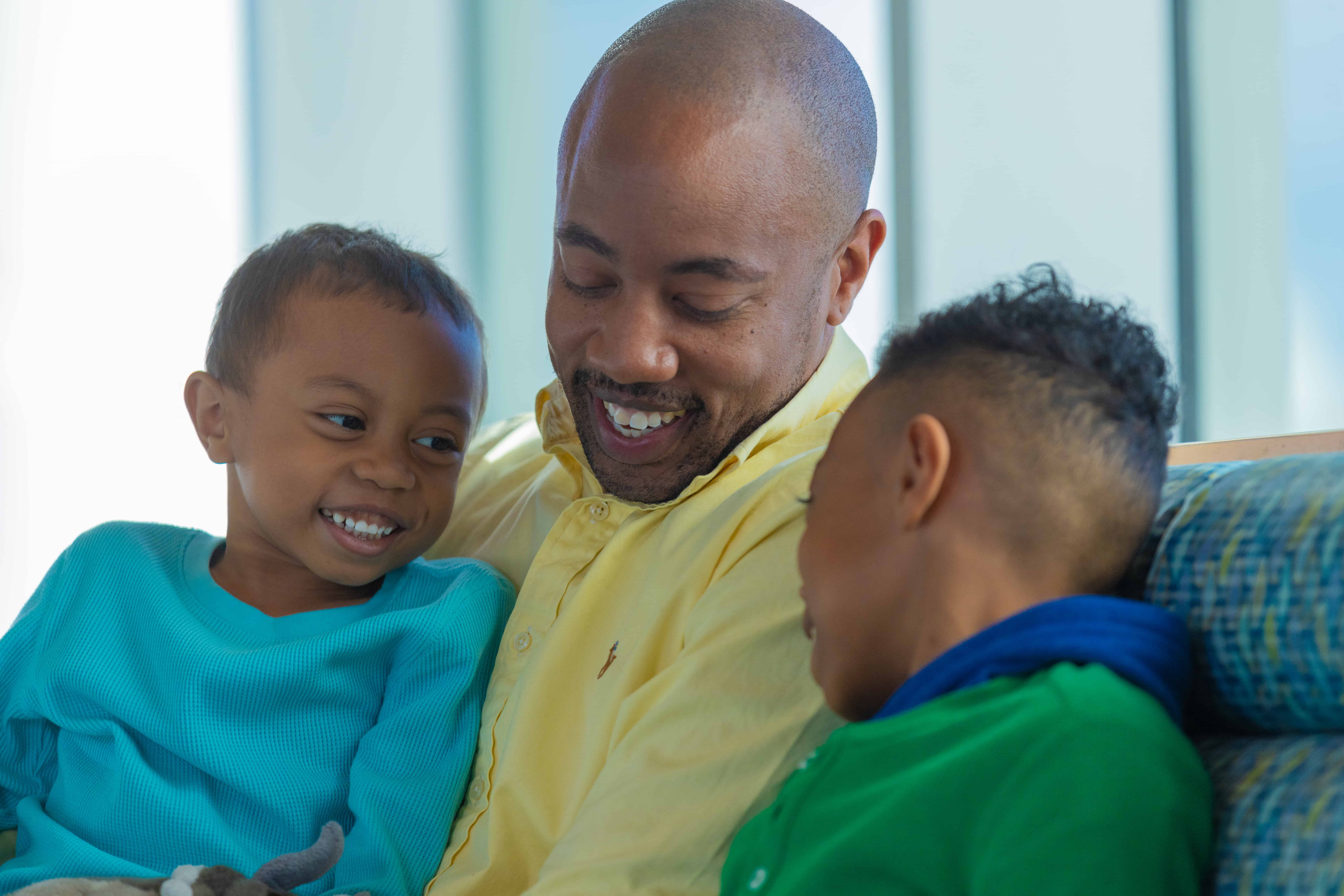 A father in a yellow shirt smiles while his two children sit next to him smiling and looking happy and playful.