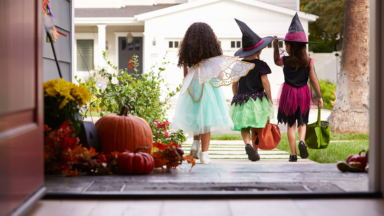 Three kids go trick-or-treating as a fairy with light blue princess dress and gold wings, a witch with black shirt, green skirt and black witch hat, and a witch in a black shirt, pink skirt and black witch hat.