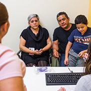 A woman in a black short sleeve sweater sits next to a man in a black t-shirt who has a boy in a blue shirt sitting on his lap while they talk to a woman sitting at a white desk with a black computer keyboard.