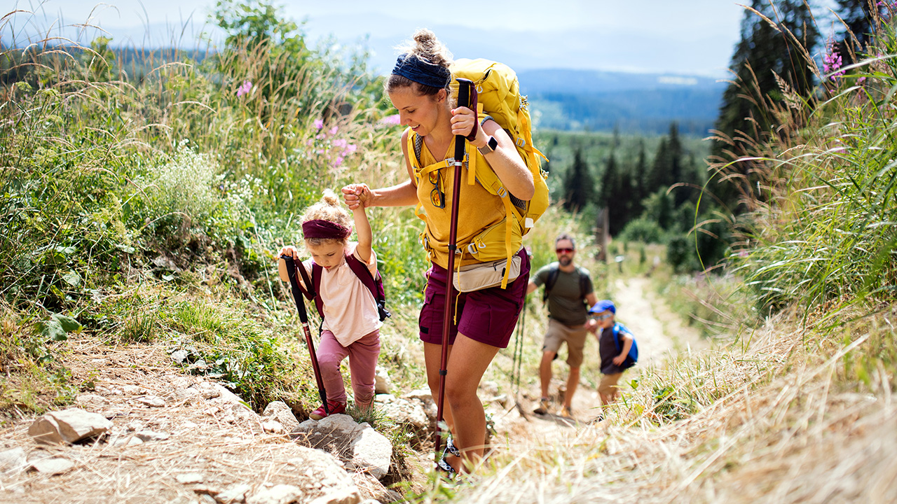 A mom helps a young girl up a rocky hiking trail, with a dad and young boy hiking in the background.