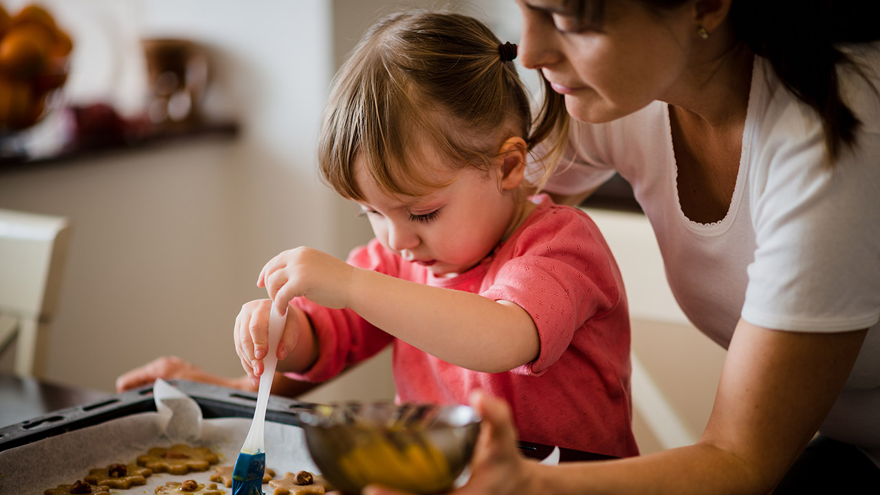 A young girl wearing a pink sweater with her sleeves rolled up paints a cookie on a baking sheet while a woman in a white shirt stands over her holding a silver bowl.