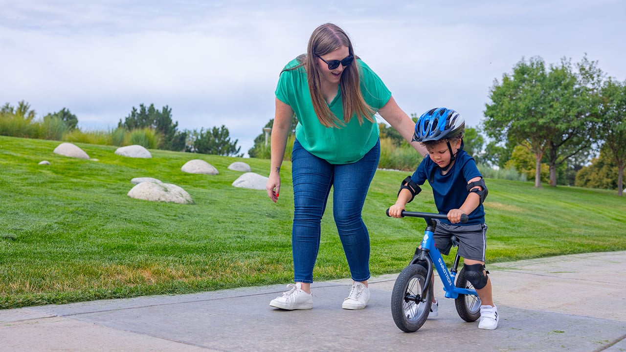  A parent with long hair, a green shirt and jeans leans over and guides her son who is on a blue bike with a blue shirt and blue helmet.