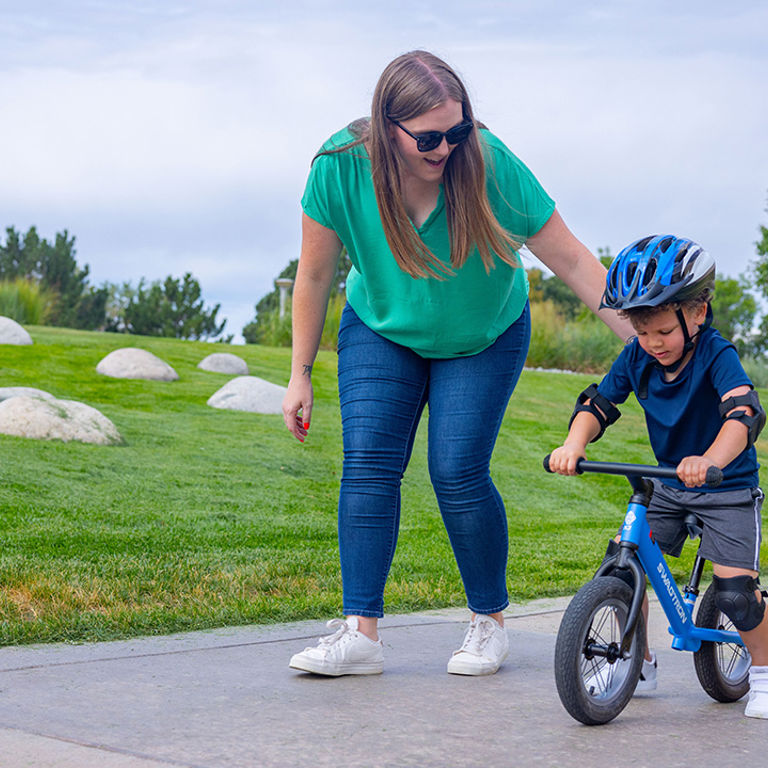  A parent with long hair, a green shirt and jeans leans over and guides her son who is on a blue bike with a blue shirt and blue helmet.