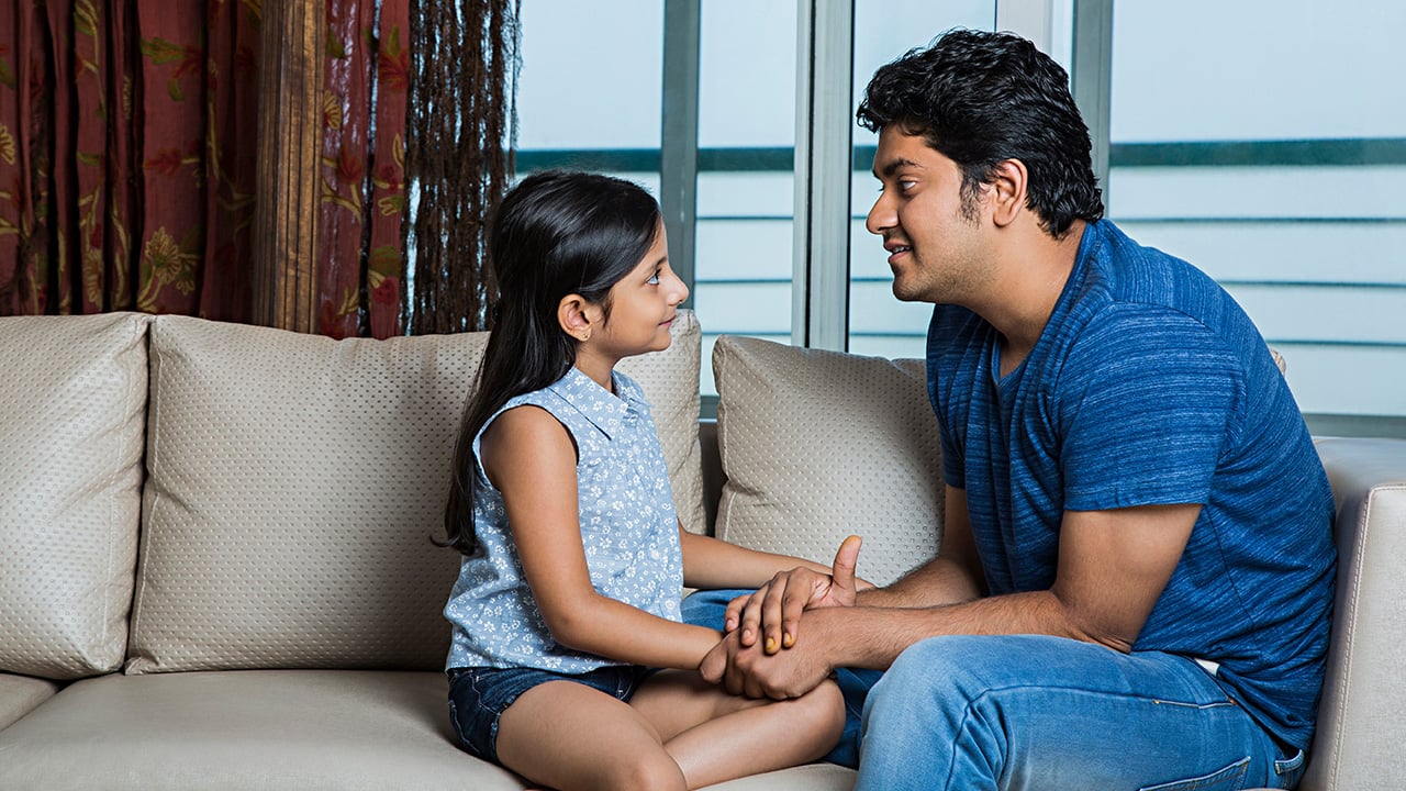 A father and daughter sit closely on a couch and talk.