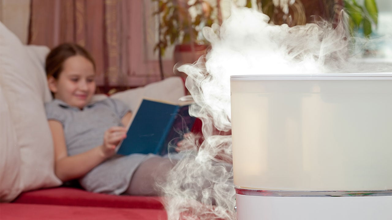 A white humidifier has a smokey vapor coming out of it while a young girl reads a blue book on a red bed with white pillows in the background.