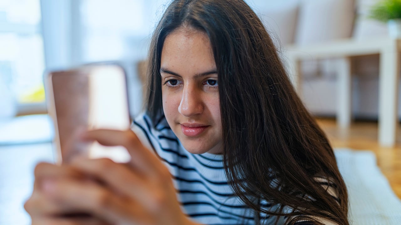 An adolescent girl with brown hair lies on her belly on the couch playing on her smartphone.