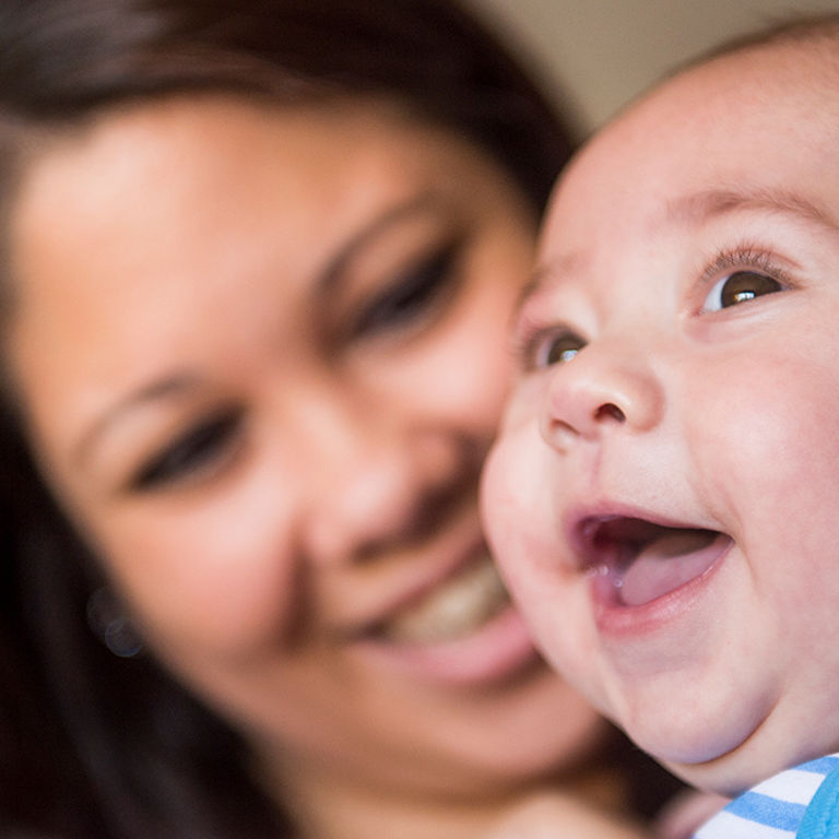 A closeup of a woman with long brown hair smiling at a happy baby wearing a blue and white stripe shirt.