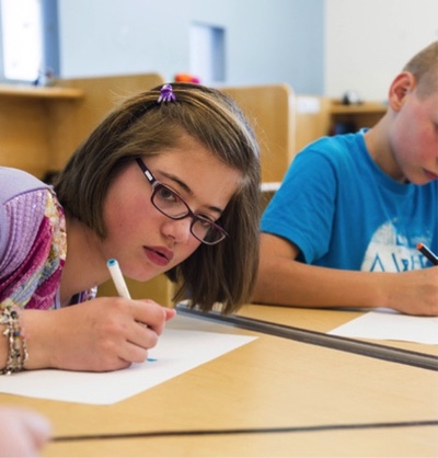 A girl with glasses writes on a piece of paper at a desk.