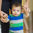 Toddler boy with brown hair walking as he holds on to his mom's hands.