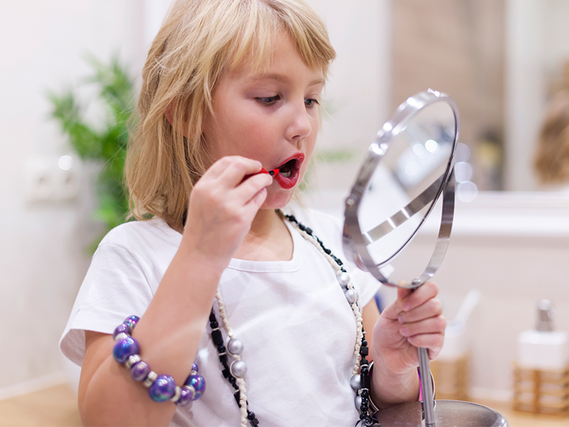 A girl wearing her mom's costume jewelry puts on lipstick while holding a vanity mirror.