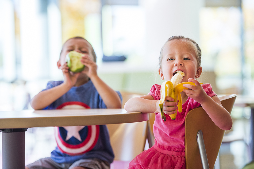 A girl in a pink dress sits at a light wood table eating a banana with a boy in a blue Captain America shirt, who is eating a green apple.