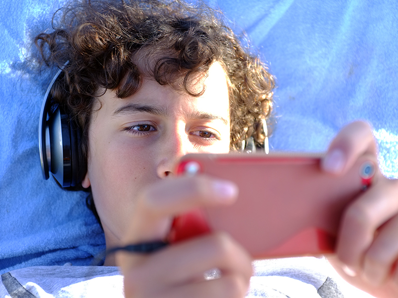 A teenage boy with brown, curly hair lies down watching his phone with big headphones on.