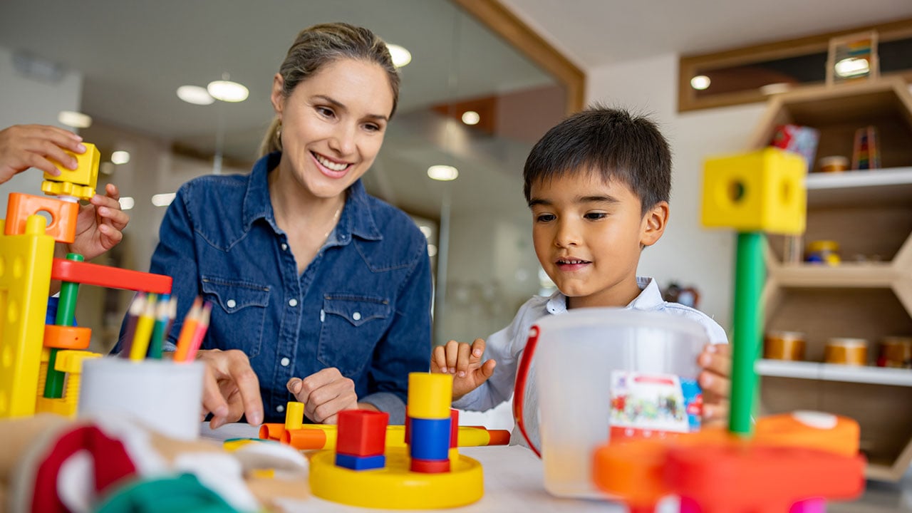 A woman watches as a young child work with bright-colored blocks at a table.