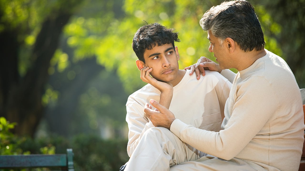 An adolescent boy and his father sit outside together while having a conversation.