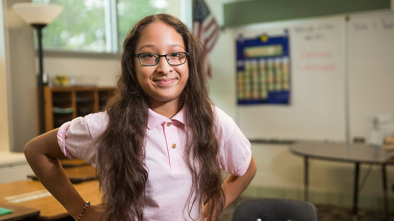 A child with long hair stands in a classroom with her hands on her hips.  