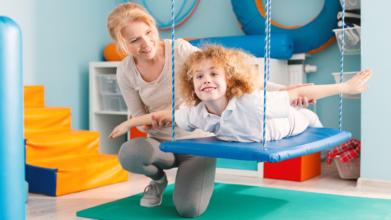 A child swings on their stomach on a sensory swing during an occupational therapy session.