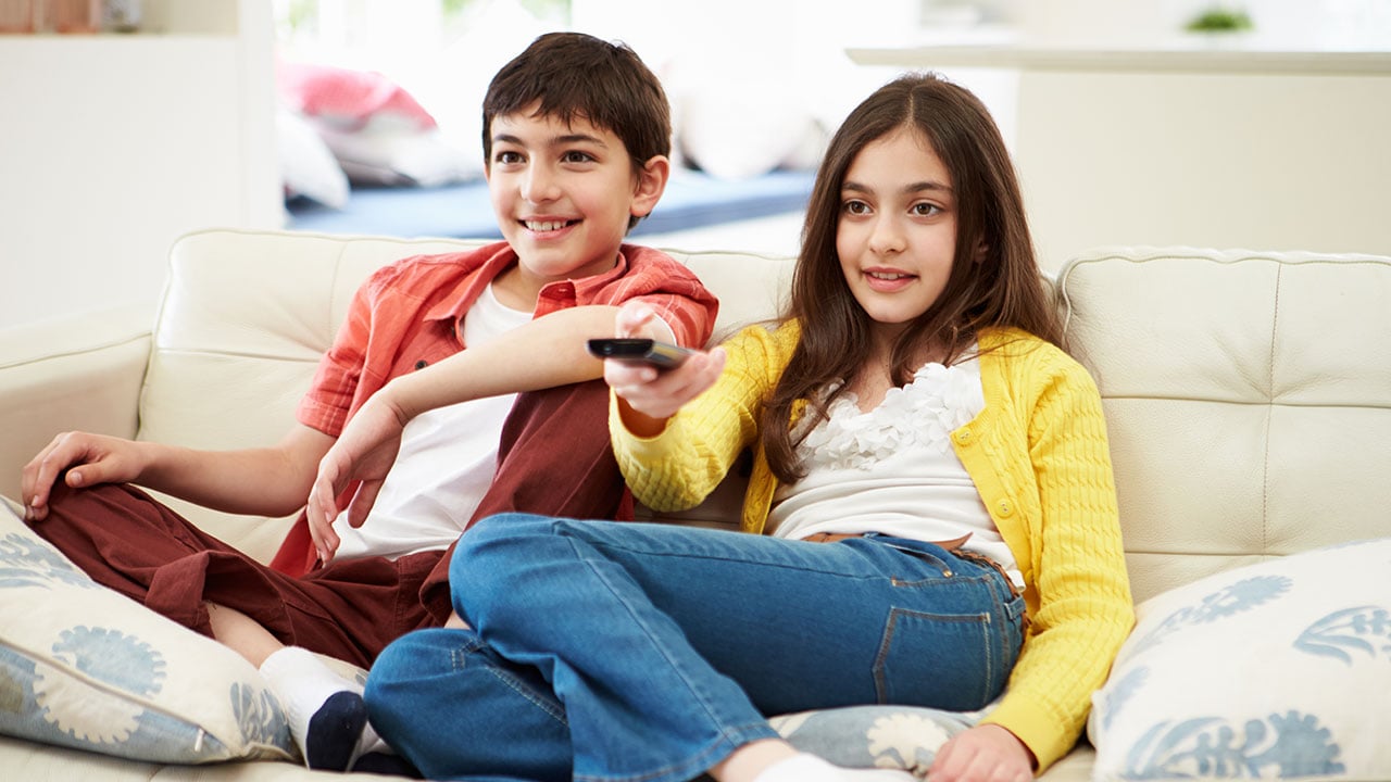 A boy and girl sit on a couch watching TV.