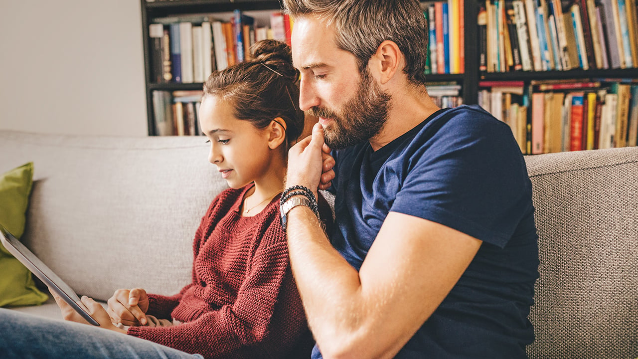 A teenage girl and her dad sit on a couch reading a tablet together.