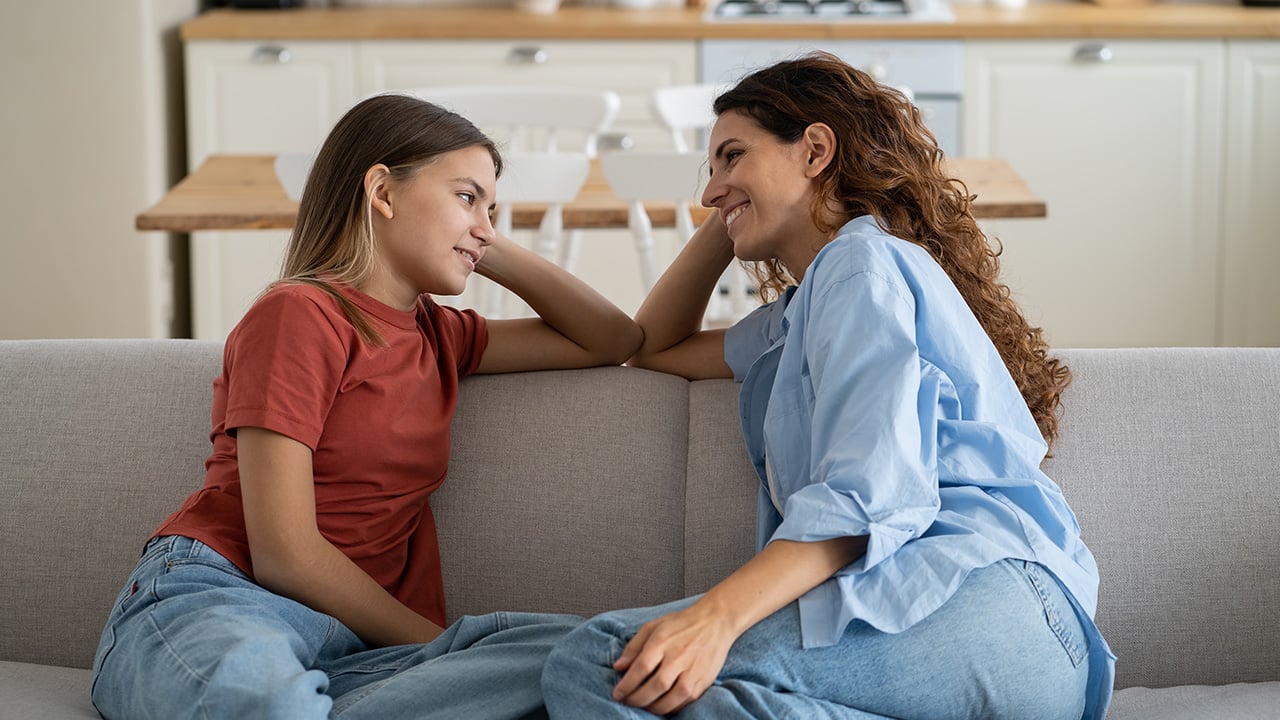 A teen girl and her mom sit on a couch smiling at each other.