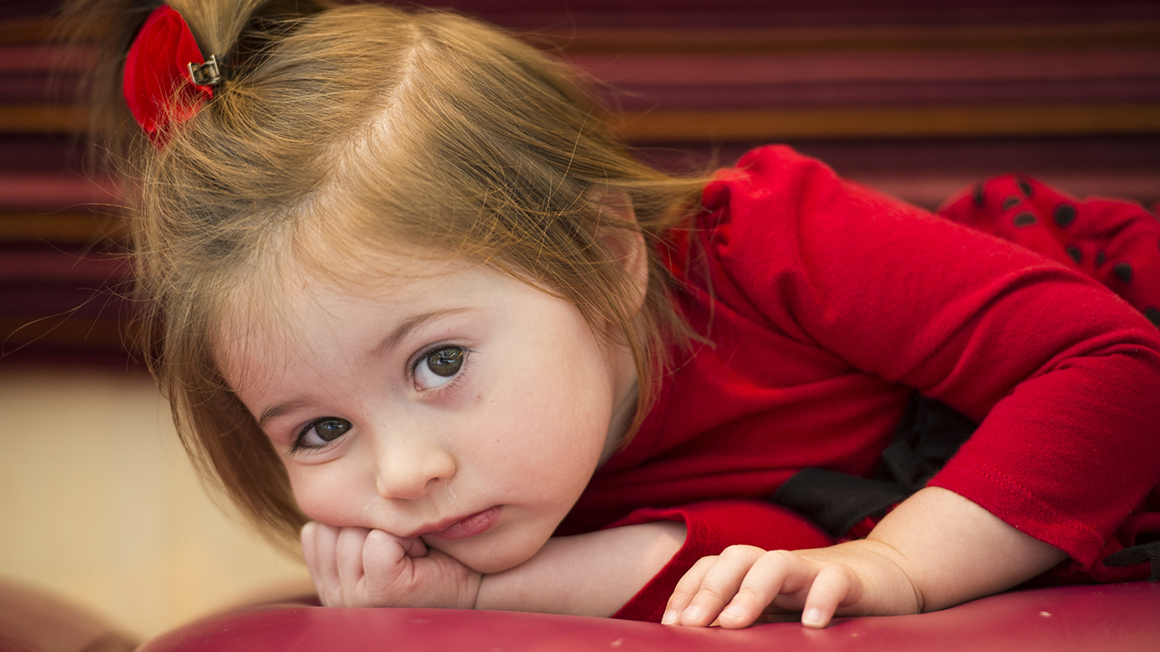 A closeup of a toddler girl with brown hair pulled back in a red bow, wearing a red dress and lying on the ground.