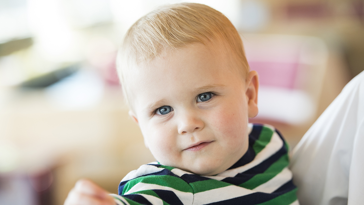 A toddler boy with blond hair and wearing a green, navy and white striped shirt is being held.