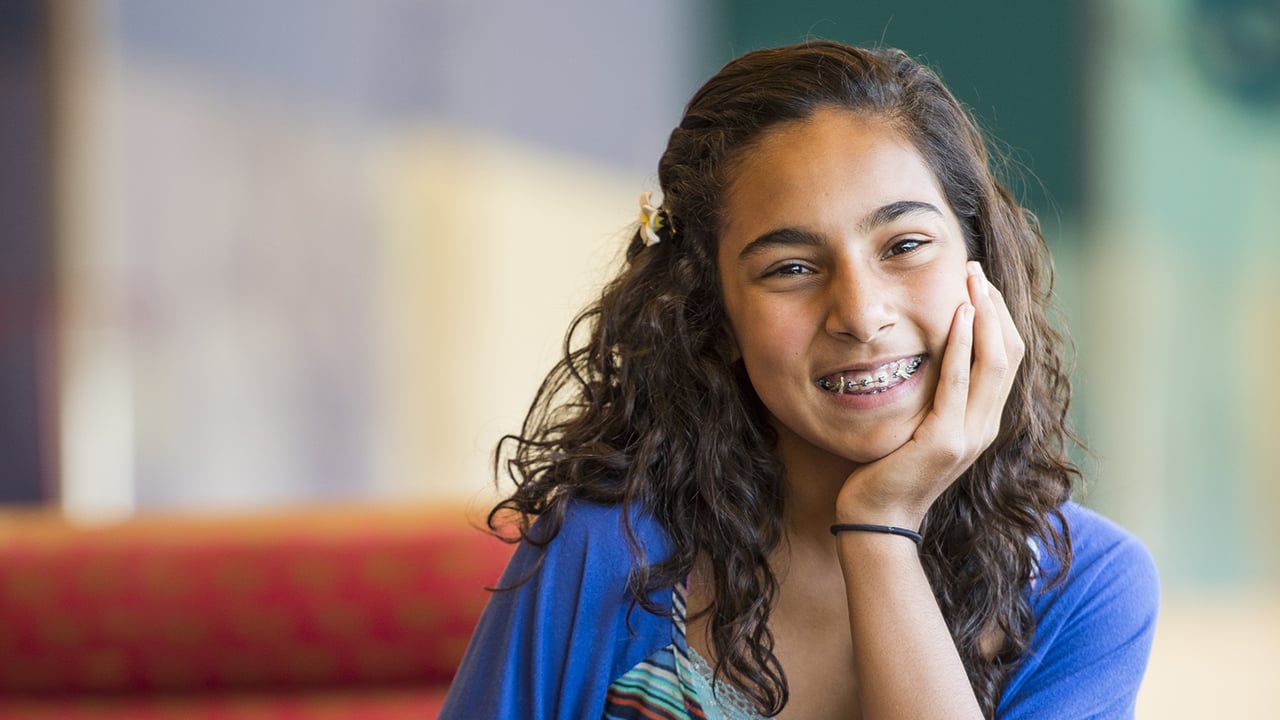 A teenage girl with long brown curly hair and braces smiles with her head resting on her hand. She is wearing a blue, green, orange, black and white striped shirt with a bright blue cardigan sweater over it.