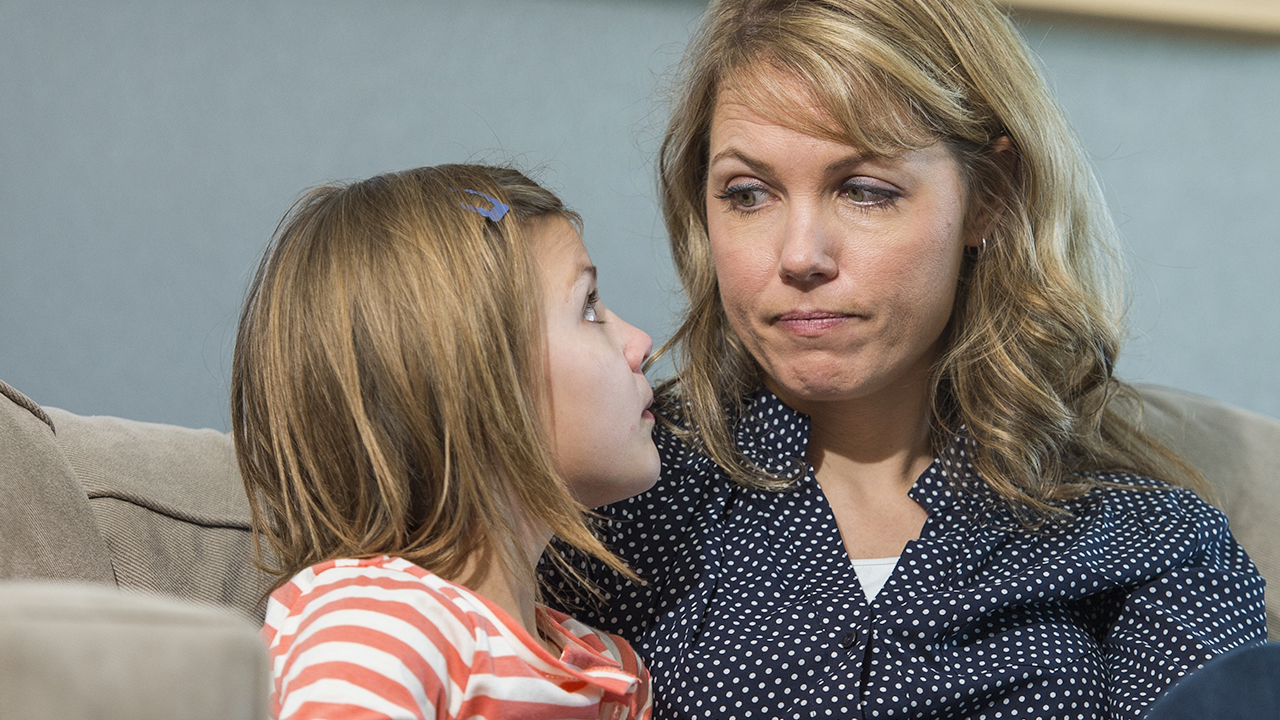 A woman with blonde hair and wearing a blue shirt with small polka-dots gives a disapproving look to a school-age girl wearing an orange and white striped shirt.