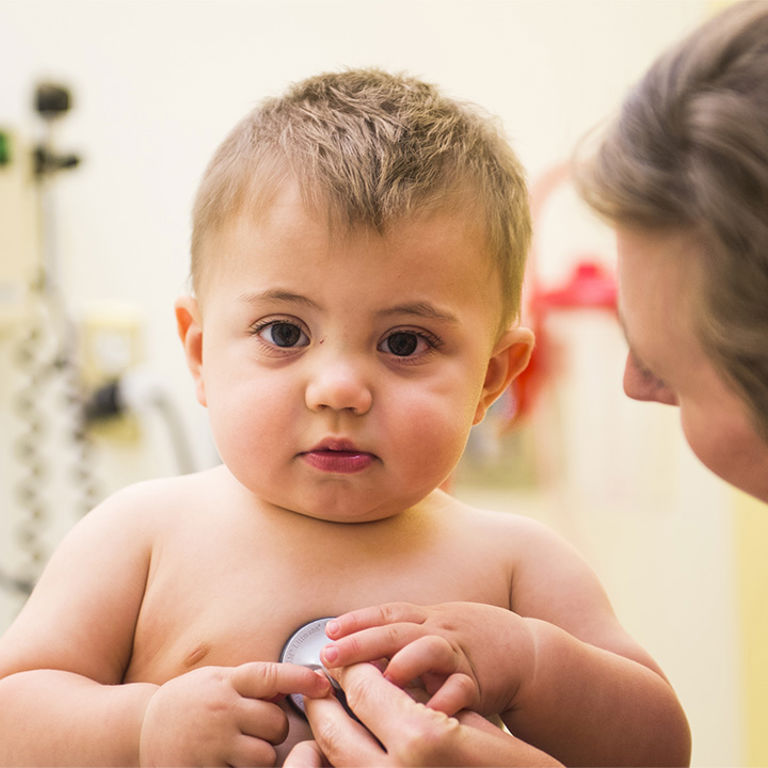 A woman with long brown hair uses a stethoscope to listen to an older baby's heart.