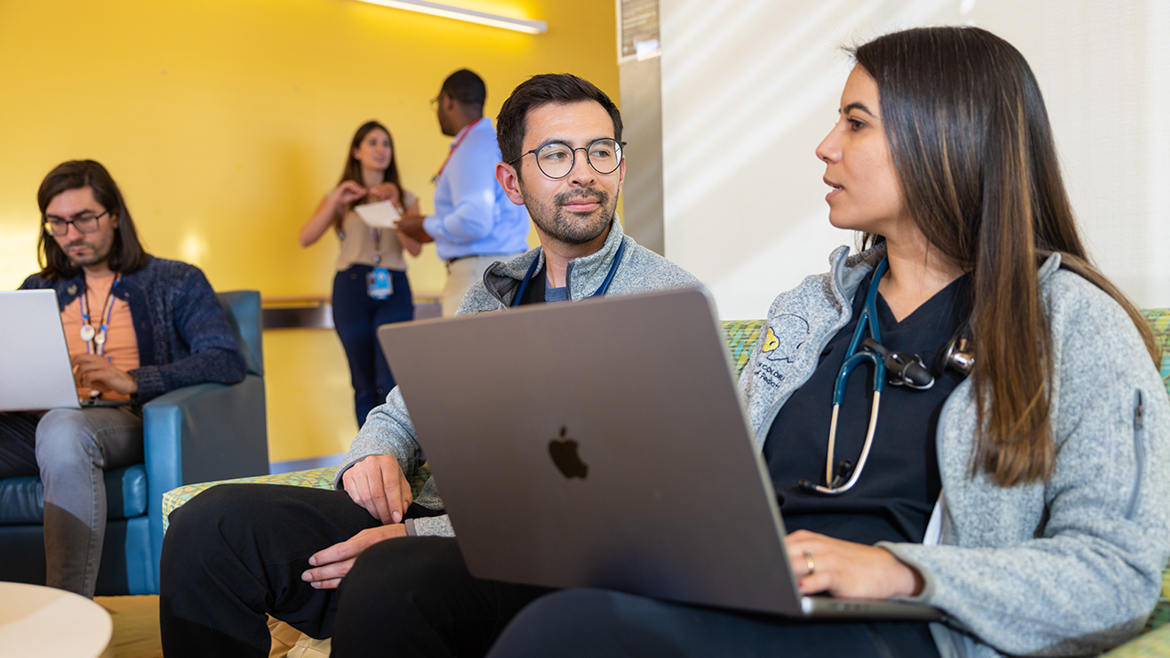 A woman and a man are seated, focused on a laptop screen and talking.