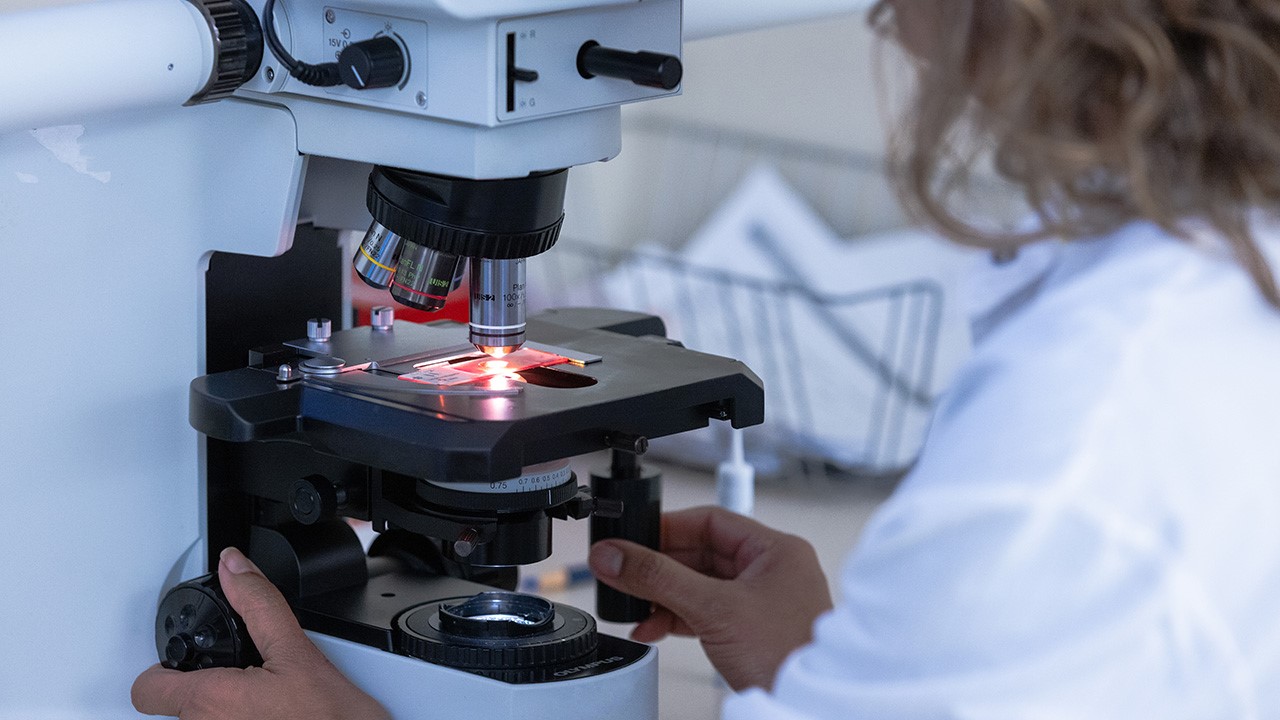 A researcher in a white lab coat looks at a slide under a microscope.