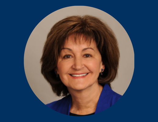 A woman with short brown hair and a blue shirt smiles at the camera. Her face is in a gray circle over a dark blue background.