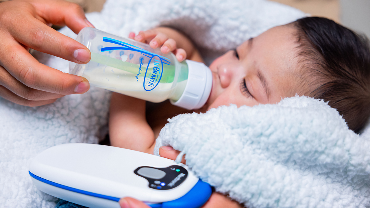 A baby wrapped in a white fuzzy blanket drinks milk from a bottle that is held by an adult hand. A small machine is next to the baby.