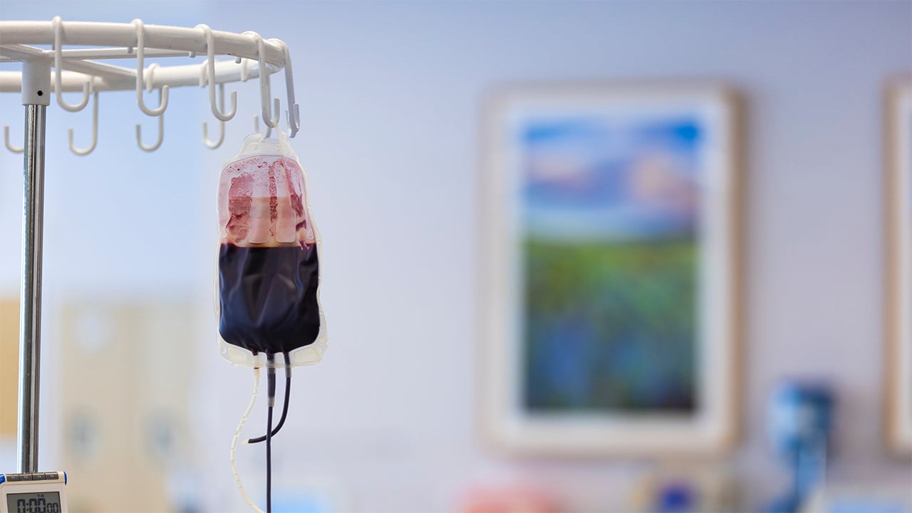 A half full blood transfusion bag hangs off a metal hook in a hospital.