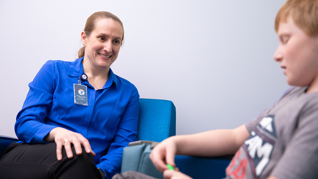 A mental health provider wearing a blue shirt sits in a chair and talks with a young patient. 