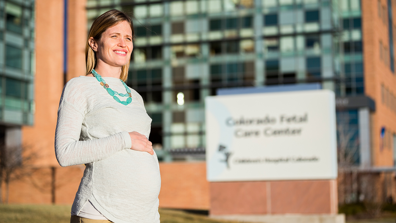 A pregnant woman in a gray shirt with her hand on her belly stands in front of the Colorado Fetal Care Center sign outside of Children’s Hospital Colorado.