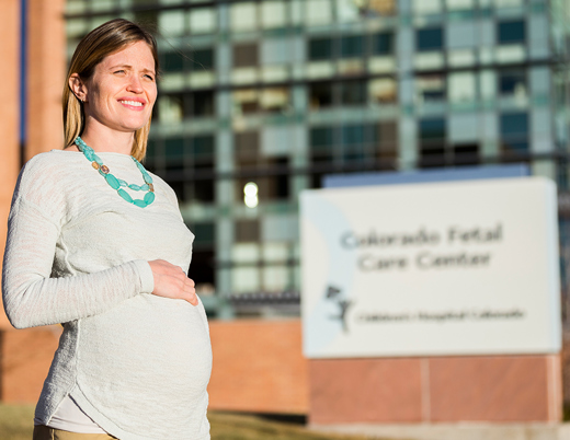 A pregnant woman in a gray shirt with her hand on her belly stands in front of the Colorado Fetal Care Center sign outside of Children’s Hospital Colorado.