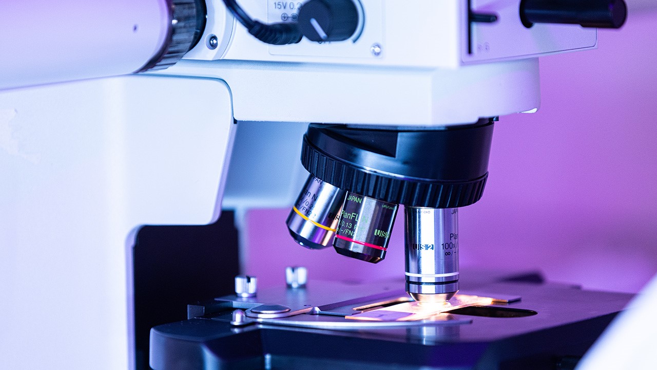 Microscope with multiple objective lenses focused on a slide, lit brightly in a dimly lit room.