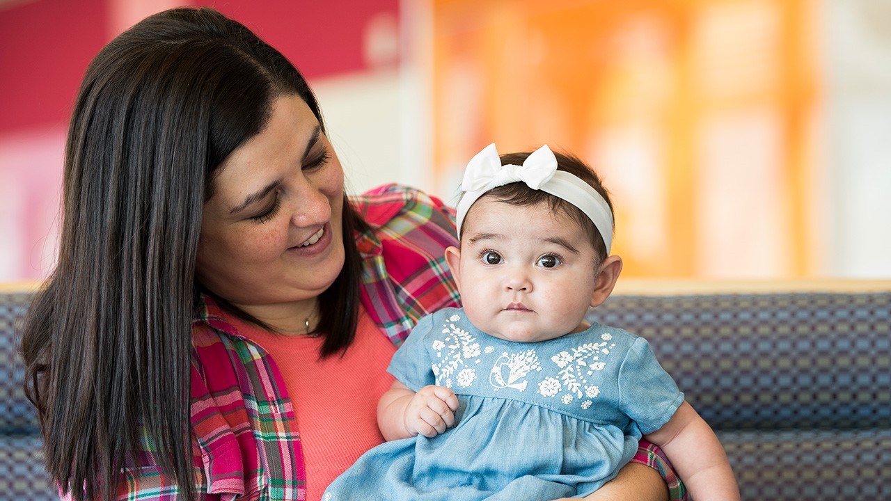 Adult holding a baby seated on a couch, both looking at something off-camera. The adult is smiling at the baby, and the baby is wearing a denim dress and a white headband.