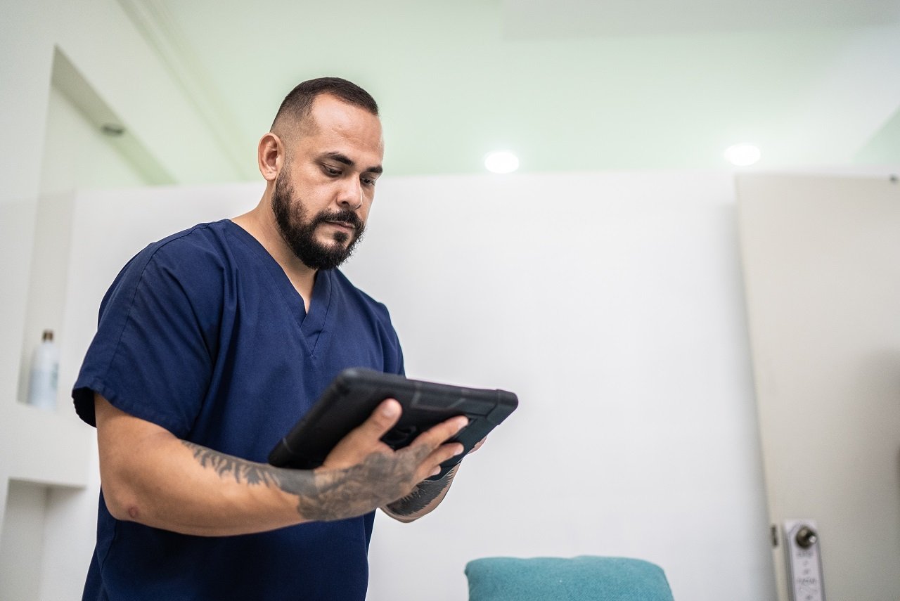 A male healthcare provider wearing dark blue scrubs stands in a hospital room looking at an iPad screen.