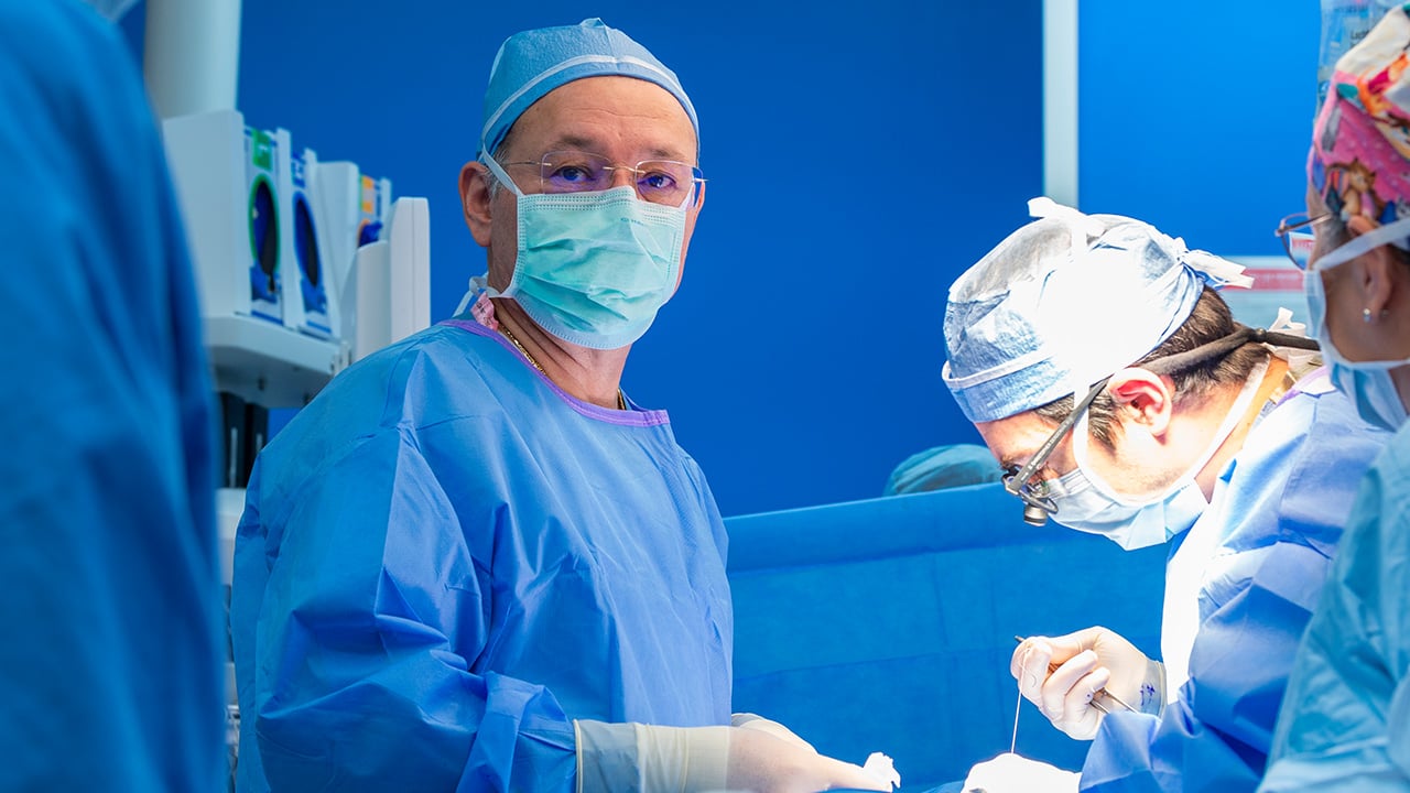 A man in scrubs, a mask and gloves in a surgery room looks at the camera while another person in scrubs holds surgical tools.