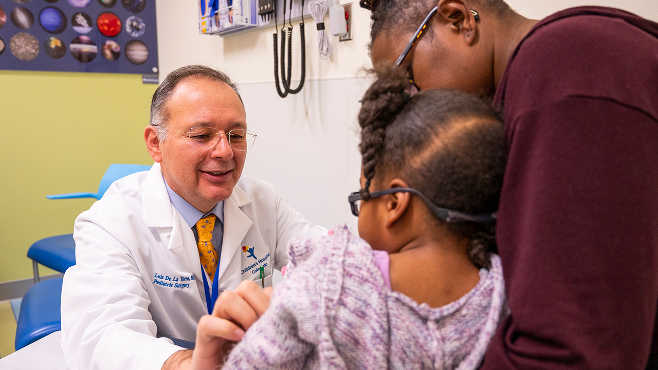 Dr. Luis De La Torre of Children’s Hospital Colorado examines a pediatric patient being held by their caregiver in a hospital exam room.